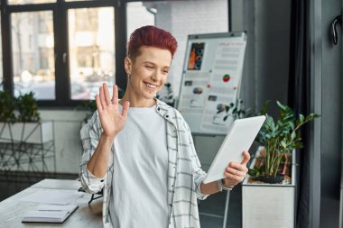 happy bigender manager waving hand during video call on digital tablet in contemporary office clipart