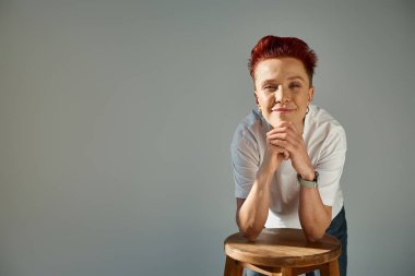 Beyaz tişörtlü pozitif çift cinsiyetli biri güneş ışığında dışkıyla poz veriyor ve gri kameraya bakıyor.