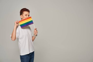 Beyaz tişörtlü kızıl saçlı eşcinsel manken. Yüzünün yanında küçük LGBT ile poz veriyor. Gri renkli kameraya bakıyor.