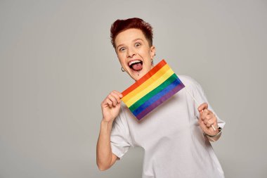 Heyecanlı tuhaf model, piercing takıyor, küçük LGBT düz duruyor ve gri arka planda dilini dışarı çıkarıyor.