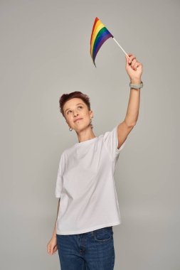 redhead queer person in white t-shirt holding small LGBT flag in raised hand while standing on grey clipart