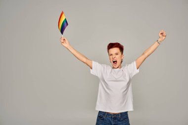 Beyaz tişörtlü, heyecanlı, küçük LGBT bayrağıyla dikilen ve gri arka planda çığlık atan tuhaf insan.