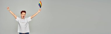 Beyaz tişörtlü, heyecanlı, küçük LGBT bayrağıyla dikilen ve gri pankartta bağıran tuhaf insan.