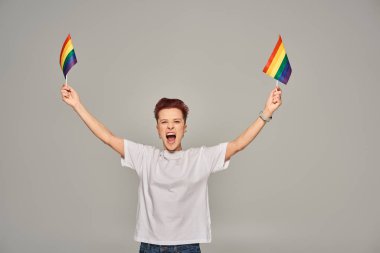 Beyaz tişörtlü, heyecanlı, küçük LGBT bayrağıyla dikilen ve gri arka planda çığlık atan tuhaf insan.