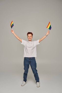 Beyaz tişörtlü ve kot pantolonlu neşeli tuhaf insan gri ellerde küçük LGBT bayraklarıyla poz veriyor.