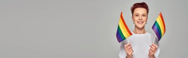 Beyaz tişörtlü neşeli tuhaf insan küçük LGBT bayrakları tutuyor ve gri pankarttaki kameraya bakıyor.