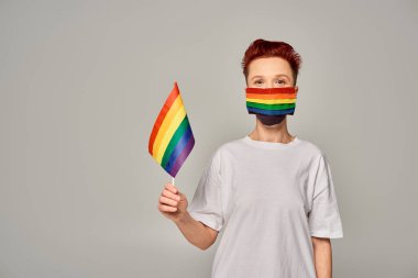 Gri tişörtlü ve gökkuşağı renkli kırmızı saçlı tuhaf adam. Üzerinde küçük LGBT bayrağı olan tıbbi maske.