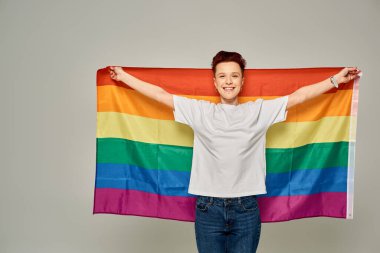 Beyaz tişörtlü neşeli kızıl saçlı adam gri arka planda LGBT bayrağıyla dikiliyor.