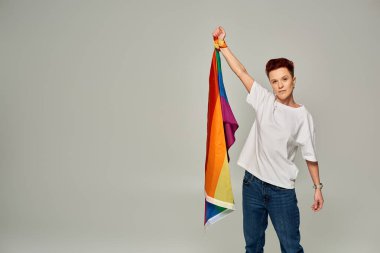 Beyaz tişörtlü ve kot pantolonlu ciddi bir kızıl saçlı. Gri arka planda LGBT bayrağı var.