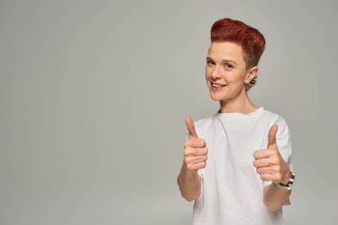 happy redhead non-binary person in white t-shirt showing thumbs and smiling at camera on grey clipart