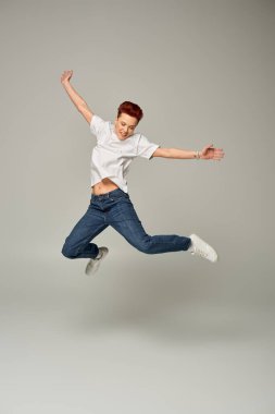 full length of happy non-binary person in white t-shirt and jeans levitating on grey backdrop clipart