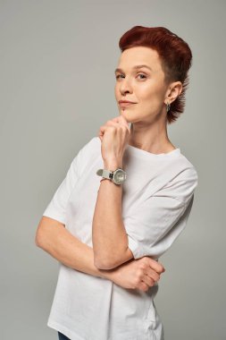 thoughtful redhead non-binary person holding hand near chin and looking at camera on grey backdrop clipart