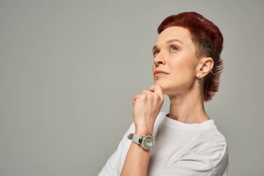 thoughtful redhead non-binary person holding hand near chin and looking away on grey backdrop clipart