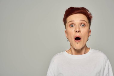 shocked redhead queer person in white t-shirt standing with open mouth and looking at camera on grey clipart