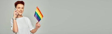 Küçük LGBT bayrağını tutan mutlu kızıl saçlı kişi ve gri pankartta akıllı telefonla konuşuyor.