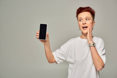 Beyaz tişörtlü, beyaz saçlı, beyaz tişörtlü, beyaz ekranlı akıllı telefon gösteren, şaşkın kızıl saçlı adam.