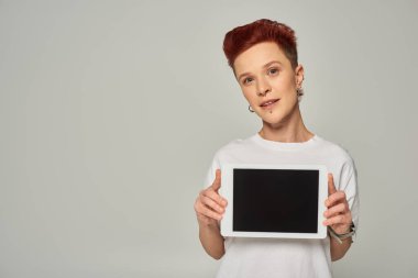 redhead queer person in white t-shirt showing digital tablet with blank screen on grey backdrop clipart