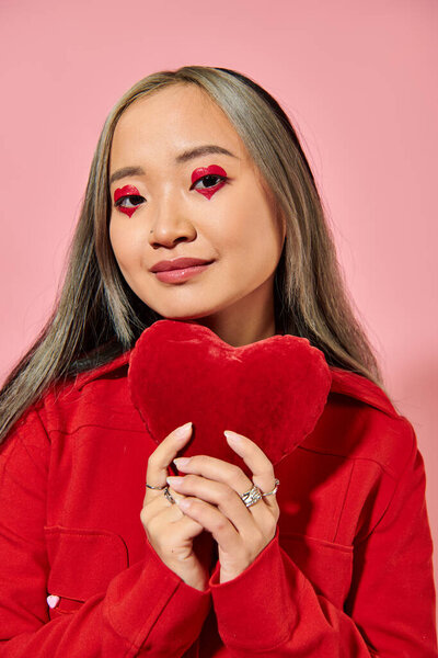 Valentines day, cheerful asian woman with vibrant eye makeup holding red heart on pink backdrop