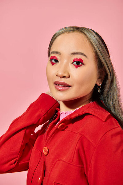 Valentines day concept, pretty asian young woman with heart eye makeup posing in red jacket on pink
