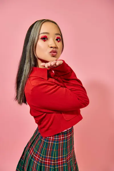 stock image asian young woman in red jacket showing air kiss gesture on pink background, heart shape eye makeup