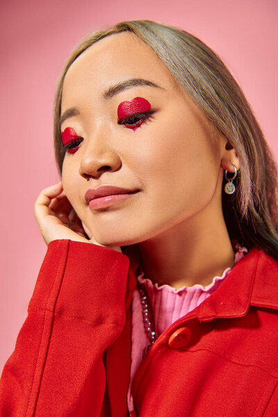 portrait of pensive asian young woman with heart shape eye makeup looking away on pink background