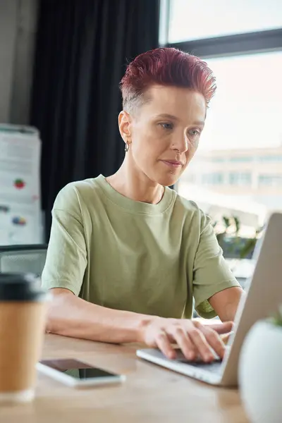 stock image stylish queer person working at laptop near blurred smartphone and coffee to go in modern office