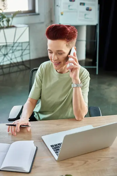 non-binary business person talking on mobile phone near laptop at workplace in modern office