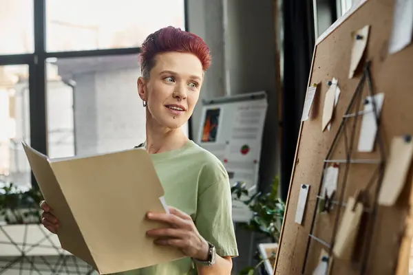 smiling queer person with documents looking at corkboard with paper notes in modern office