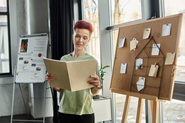 smiling queer person looking at folder with documents near corkboard with paper notes in office