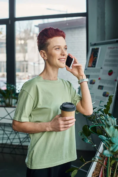 happy queer person with coffee to go in paper cup talking on mobile phone in modern office