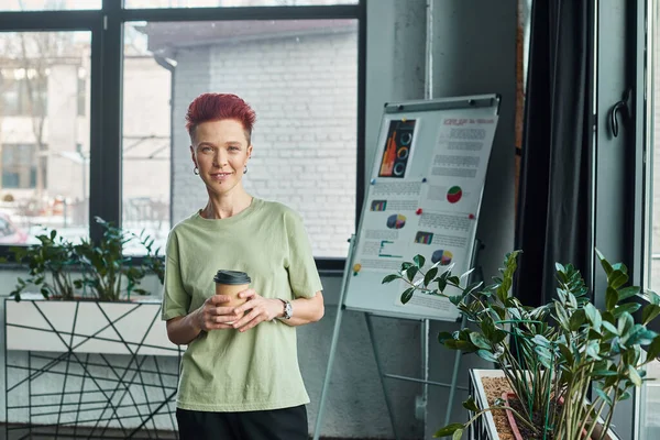 smiling non-binary person with takeaway drink in paper cup looking at camera in modern office