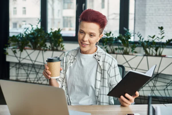 stylish non-binary person with coffee to go and notebook sitting near laptop in modern office