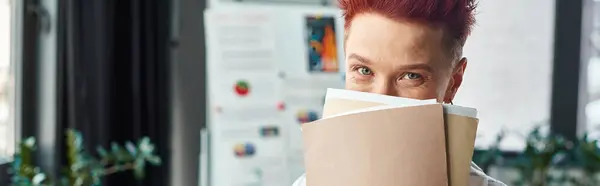 cheerful bigender person with obscuring face with folders and looking at camera in office, banner