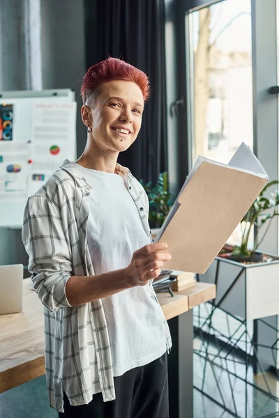 joyful bigender person in casual attire standing documents and looking at camera in modern office