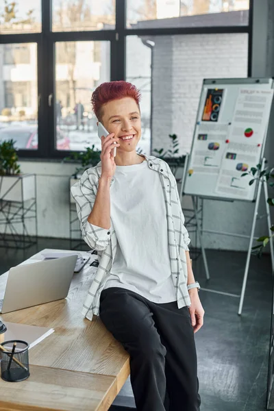 smiling bigender manager in casual attire sitting on work desk and talking on smartphone in office