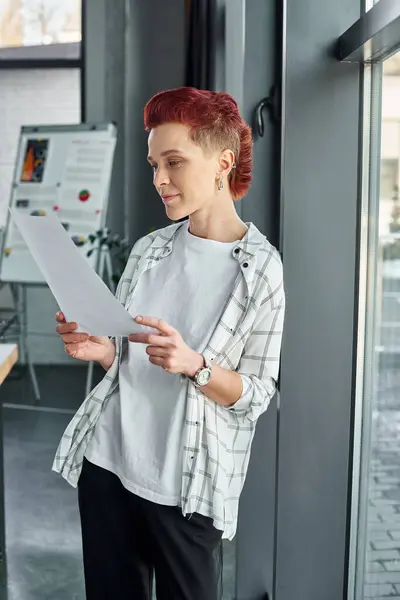thoughtful non-binary person in casual attire looking at documents while standing in modern office