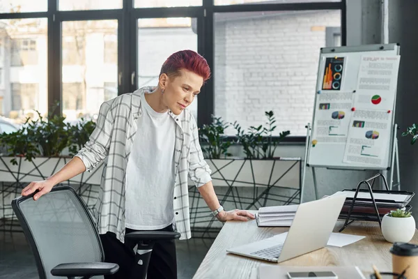 thoughtful non-binary person in casual attire standing at workplace in office and looking at laptop