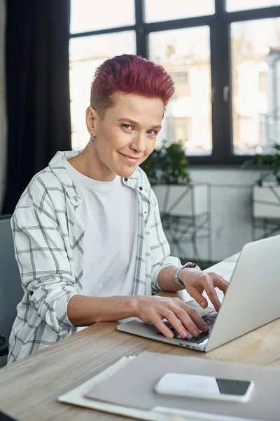 stylish queer person in casual attire smiling at camera while working on laptop in modern office