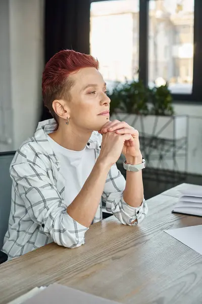 thoughtful non-binary manager with hands near chin sitting at workplace in office and looking away