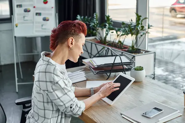 non-binary person in casual attire using digital tablet near documents and smartphone in office