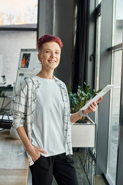 happy bigender manager in casual attire standing in office with digital tablet and looking at camera