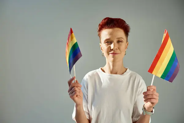 redhead non-binary person in white t-shirt standing with small rainbow colors flags on grey