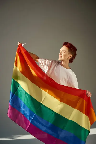 happy and unique queer person in white t-shirt posing with rainbow colors LGBT flag on grey backdrop