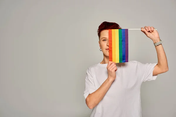 redhead queer person in white t-shirt obscuring face with small LGBT flag looking at camera on grey