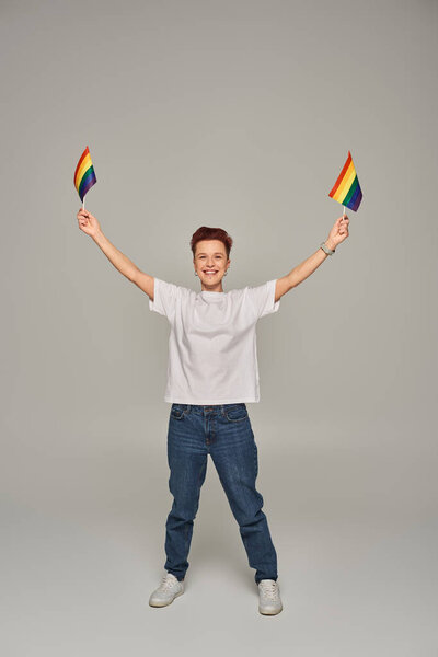 joyful queer person in white t-shirt and jeans posing with small LGBT flags in raised hands on grey