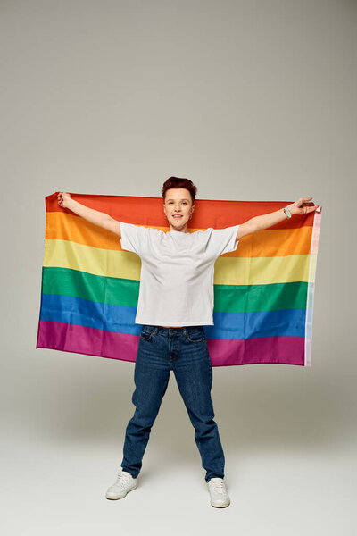 full length of redhead bigender person in white t-shirt and jeans standing with LGBT flag on grey
