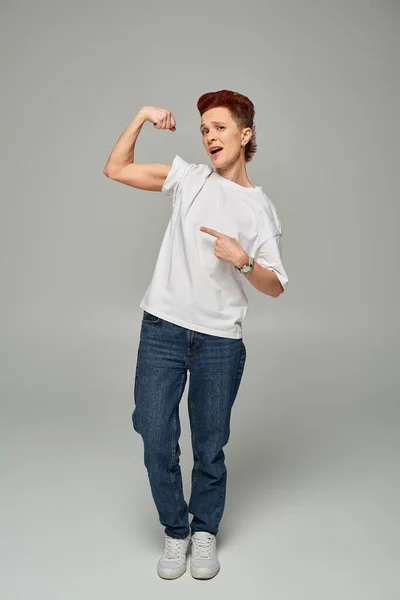 displeased non-binary person in white t-shirt showing and pointing at weak muscles on grey