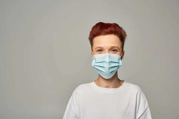 portrait of redhead queer person with happy gaze wearing medical mask and looking at camera on grey