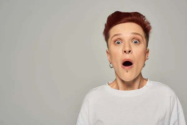 stock image shocked redhead queer person in white t-shirt standing with open mouth and looking at camera on grey