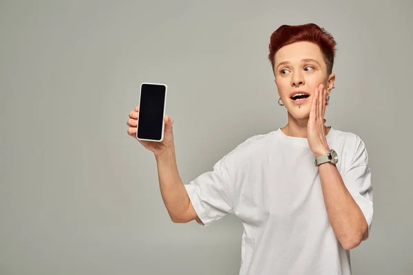 amazed redhead queer person in white t-shirt showing smartphone with blank screen on grey